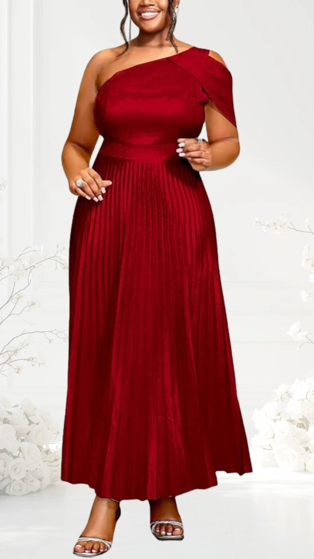A vibrant red, one-shoulder wedding guest dress features a fitted bodice that flows into a pleated skirt, creating an elegant silhouette. The dress is complemented by a subtle shoulder detail and is paired with strappy silver sandals. The setting includes soft white floral accents in the background, enhancing the overall romantic feel.