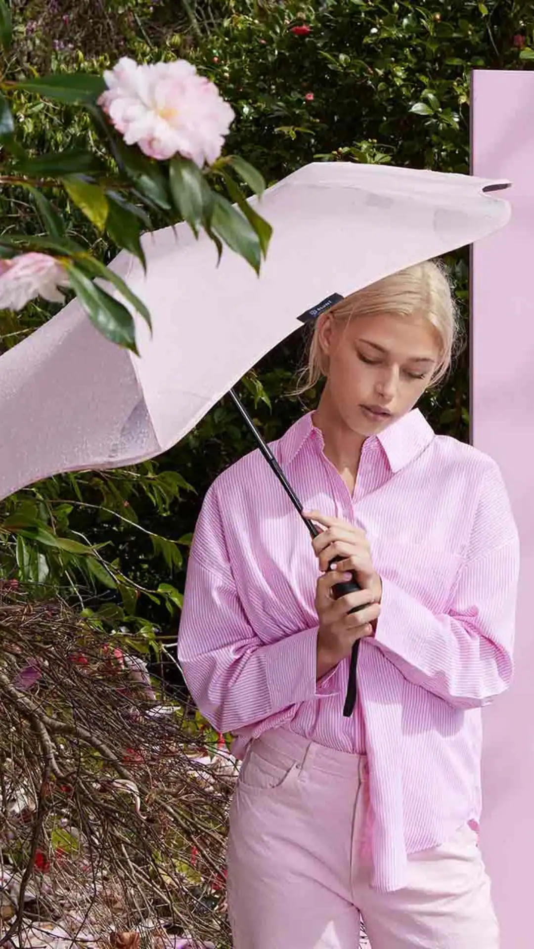This summer outfit features a stylish oversized pale pink striped button-up shirt paired with matching light pink trousers, both crafted from soft, lightweight fabric for comfortable wear during rainy days. The overall vibe is relaxed yet chic, making it perfect for casual outings or brunch, complemented by a coordinating light pink umbrella for added functionality.