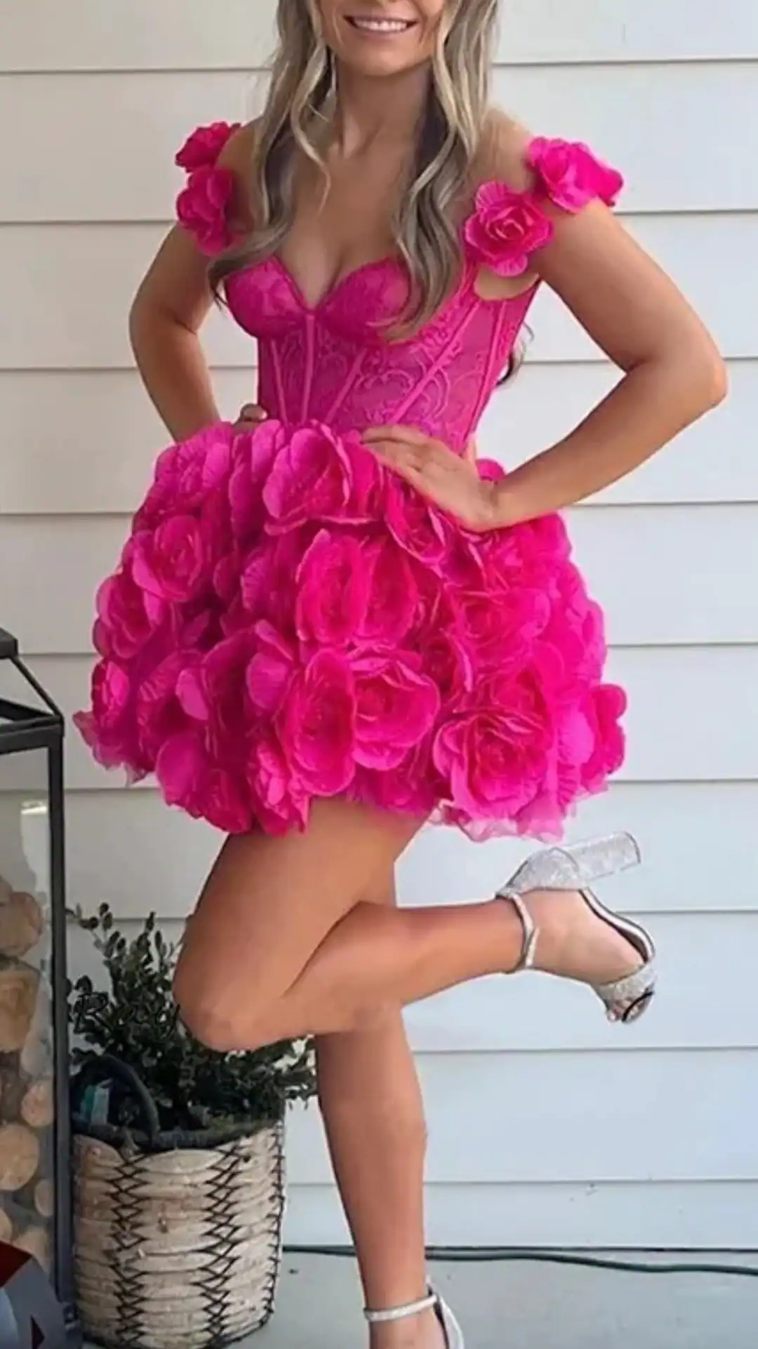 This bright pink homecoming dress features a structured corset bodice adorned with intricate detailing, complementing the voluminous skirt made of layered, oversized rose appliqués, creating an eye-catching floral effect. The style is whimsical and playful, making it perfect for a teen celebrating homecoming or a school dance.