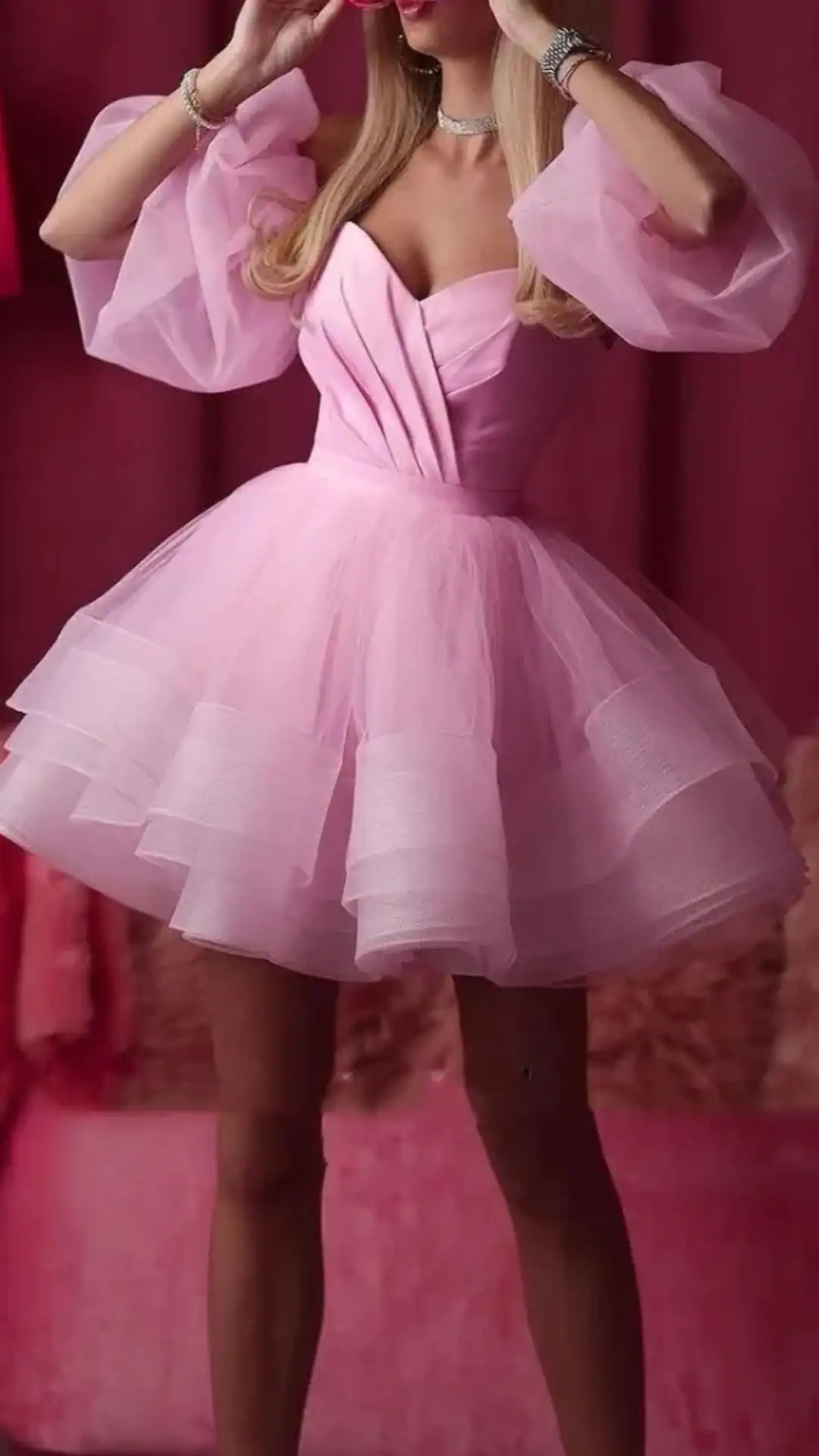 This playful homecoming dress features a soft, pastel pink tulle fabric with a structured corset-like bodice and voluminous layered skirt that exudes a youthful, fairy tale charm. The puffed sleeves add a romantic touch, making it a perfect choice for a formal event or school dance for teens.