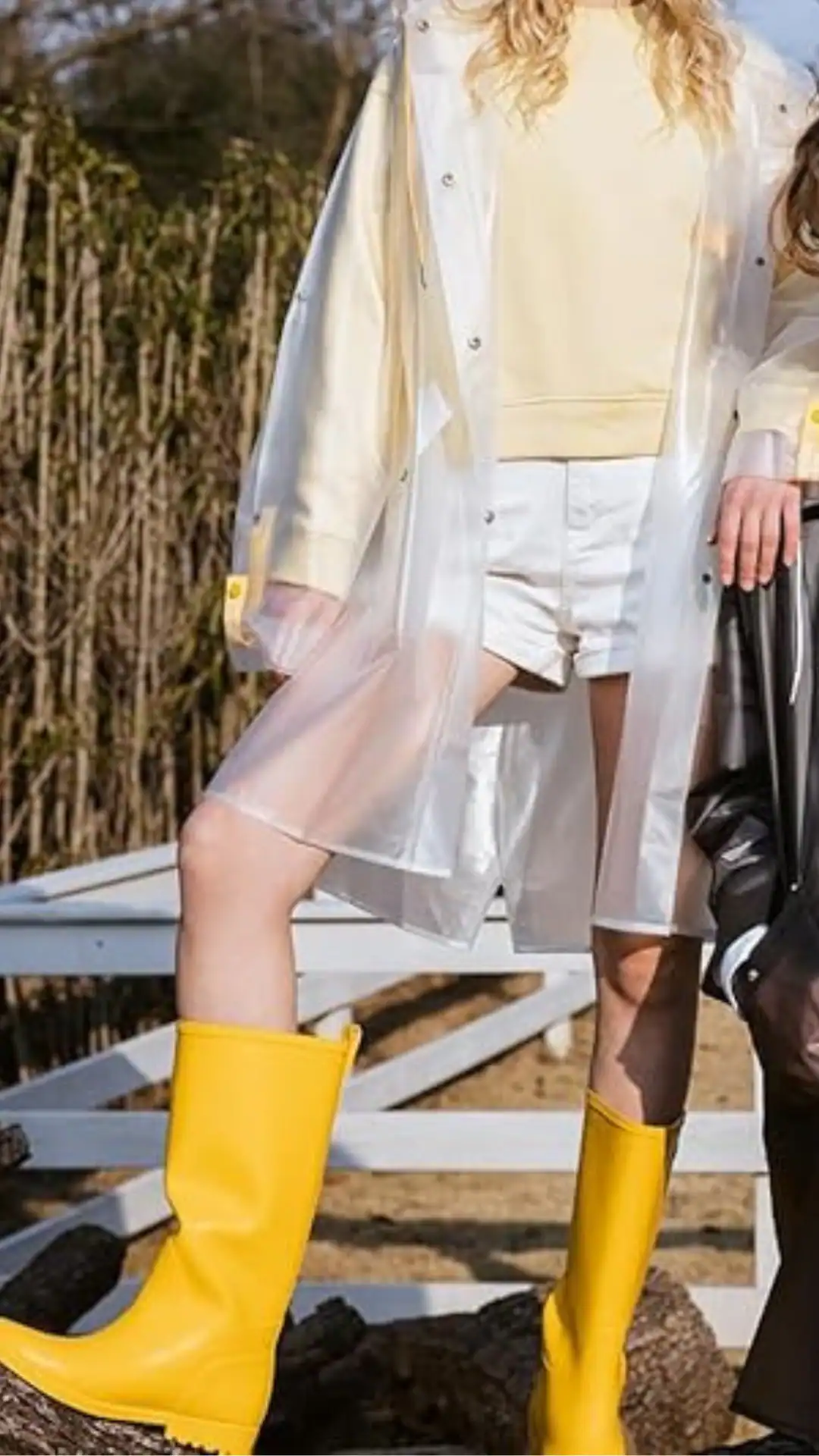 A playful summer outfit featuring a translucent, knee-length raincoat layered over a soft, pastel yellow sweatshirt and white shorts. The ensemble combines glossy textures with a vibrant color palette, highlighted by bold, knee-high yellow rain boots, exuding a cheerful and adventurous vibe, perfect for casual outings in rainy weather.