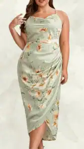 This plus-size wedding guest dress features a soft green base adorned with a delicate floral pattern in shades of peach and cream. The dress has a fitted silhouette with a draped waist design, showcasing a flattering side slit. Thin spaghetti straps complement the stylish cowl neckline, creating an elegant and feminine look perfect for special occasions.