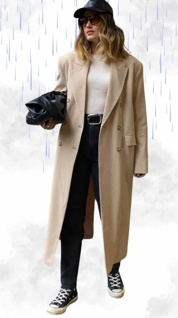 The outfit features a long beige double-breasted coat layered over a fitted white ribbed top, paired with dark high-waisted jeans for a sleek silhouette. Complementing the look are classic black Converse sneakers and a black bucket hat, while a glossy black handbag adds a chic touch, perfect for staying stylish and warm on rainy days.