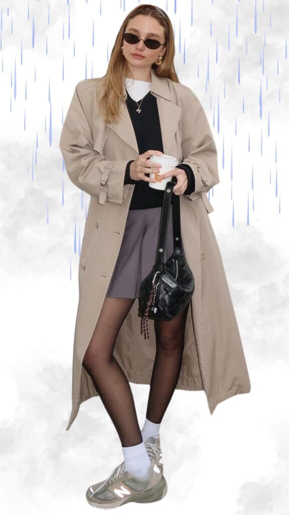 The outfit features a light beige trench coat layered over a black sweater, paired with a chic purple skirt. The look is complemented by sheer black tights, white ankle socks, and stylish gray sneakers with reflective accents. Accessories include small black sunglasses and a textured black handbag, perfect for a trendy, warm, and dry ensemble on a rainy day.