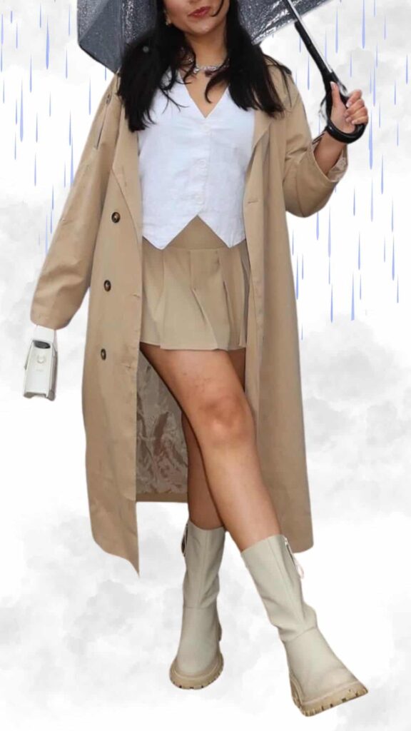 The outfit features a light beige waterproof trench coat layered over a crisp white cropped blouse and a matching beige pleated mini skirt, combining classy and trendy elements. Paired with knee-high cream rain boots that have a textured sole for stability, this ensemble is completed with a clear umbrella, ensuring style while keeping dry in rainy fall and winter weather. The overall color palette is soft and neutral, perfect for a chic, seasonal look.