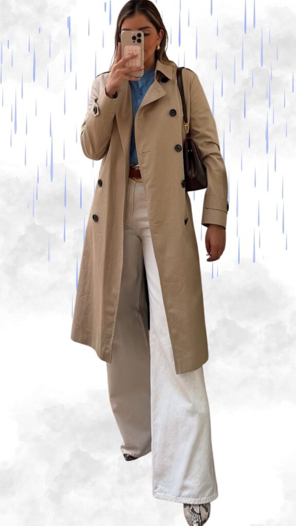 The outfit features a light beige, double-breasted trench coat made of a water-resistant material, layered over a soft blue button-up shirt. Wide-leg white pants create a chic silhouette, complemented by stylish, patterned ankle boots. Accessories include a brown leather crossbody bag and a gold belt, adding a polished touch to this elegant rainy day ensemble.