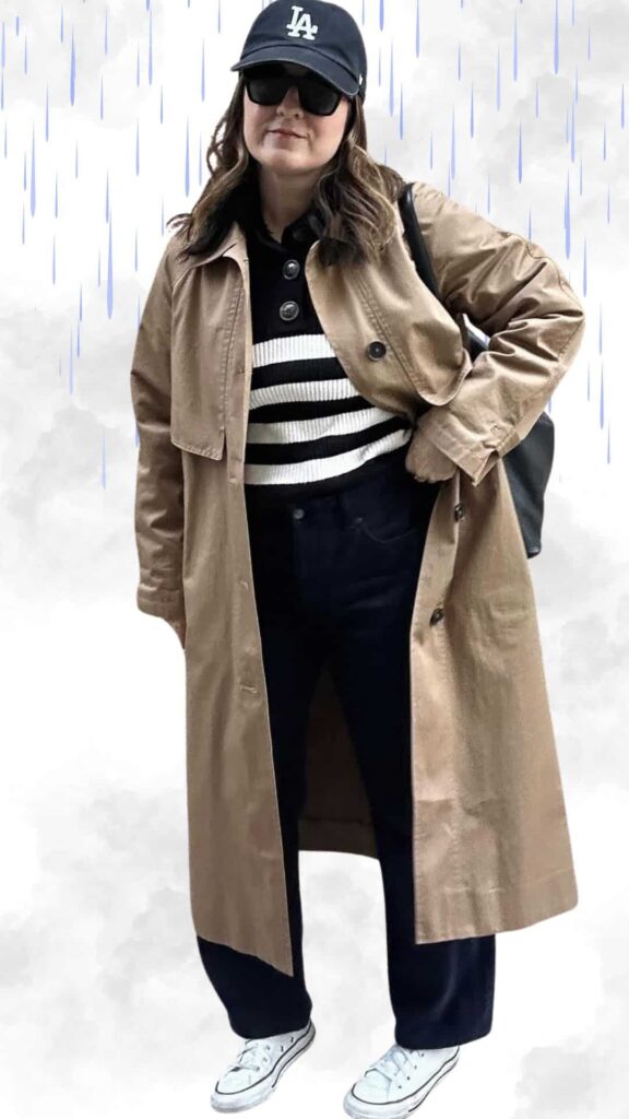 The outfit features a soft, beige trench coat layered over a black and white striped sweater, providing both warmth and a chic aesthetic. Paired with dark, relaxed-fit jeans and classic white sneakers, this rainy day look is completed with a sporty black cap and oversized sunglasses for a stylish yet practical touch. The combination of textures and colors ensures a fashionable appearance while remaining comfortable and dry in fall and winter weather.