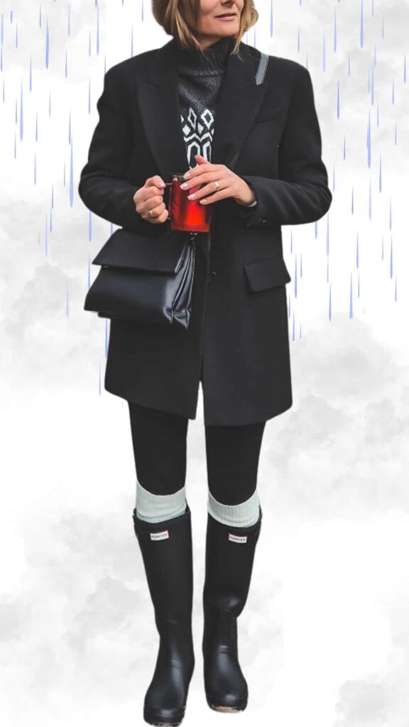 The outfit features a tailored black coat over a cozy, patterned knit sweater, paired with form-fitting black leggings for warmth. Glossy black rain boots with a ribbed cuff in light gray add a stylish touch, while a sleek black handbag and a warm drink complete the chic autumn look.