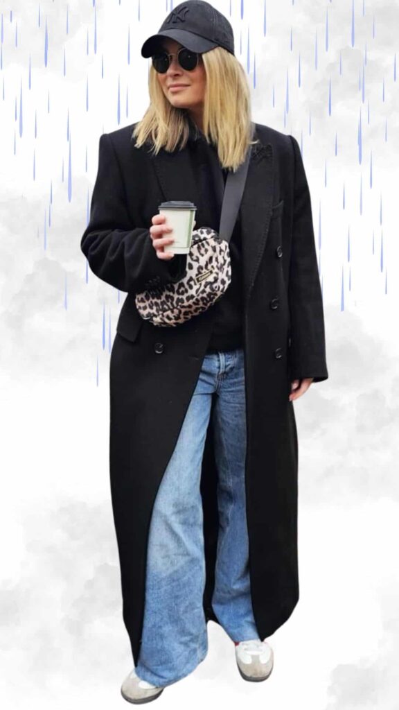 The outfit features an oversized black coat layered over a black sweater, paired with wide-leg blue denim jeans. Accessories include a black baseball cap, round sunglasses, and a spotted crossbody bag, while the shoes are stylish gray slip-on boots, all combining practicality and chic aesthetics for rainy fall and winter days.