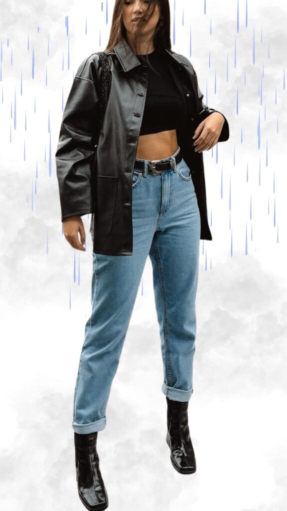The outfit features a sleek black faux leather oversized coat, paired with a fitted black crop top that highlights a minimalist look. It includes light blue high-waisted jeans with a cuffed hem, balanced by shiny black ankle boots, creating a chic and modern appearance suitable for rainy fall and winter weather. A woven black handbag adds a stylish accessory, enhancing the overall classy vibe while ensuring warmth and protection from the elements.