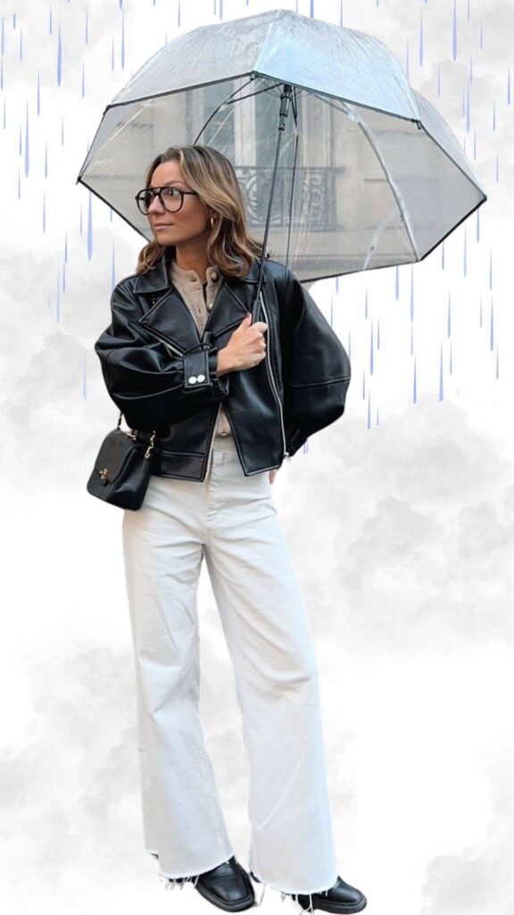 The outfit features a chic black cropped leather jacket with a structured silhouette, layered over a soft beige sweater. Paired with wide-leg white trousers that have raw hems and sleek black ankle boots, the look is completed with a clear umbrella and a small black crossbody bag, creating a stylish and practical ensemble for rainy fall and winter weather.