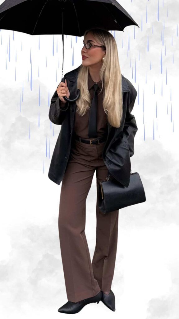 The outfit features a chic brown ensemble, including tailored wide-leg trousers and a matching shirt, layered with a sleek black waterproof coat that adds sophistication. Complemented by pointed black ankle boots and a structured black handbag, the look is completed with a classic black umbrella, making it both stylish and practical for rainy fall and winter weather.