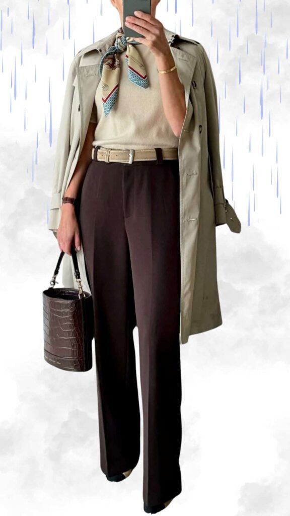 The outfit features a light beige sweater layered under a classic khaki trench coat, paired with elegant, wide-leg dark brown trousers that offer a sophisticated silhouette. A chic silk scarf adds a pop of color around the neckline, complemented by a stylish brown croc-embossed handbag and polished black ankle boots for a refined, warm, and waterproof look perfect for rainy fall and winter days.