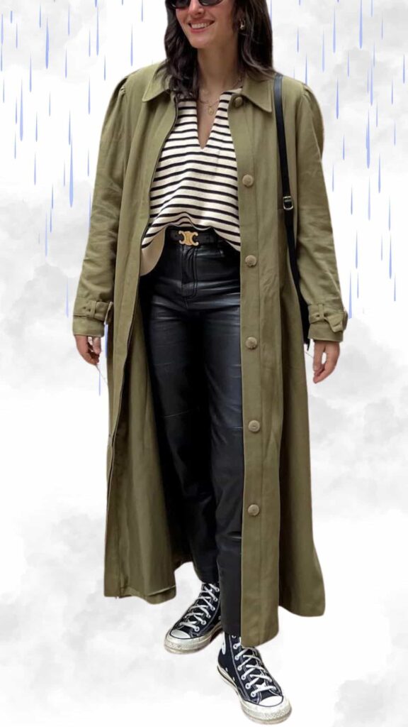 A chic outfit featuring a long olive-green trench coat with a relaxed fit, button detailing, and rolled sleeves, layered over a classic black and white striped top. The ensemble includes sleek black faux leather trousers and stylish black high-top sneakers, completing the look with a modern touch. Minimalist jewelry and sunglasses add a polished finish, perfect for staying warm and fashionable on a rainy day.