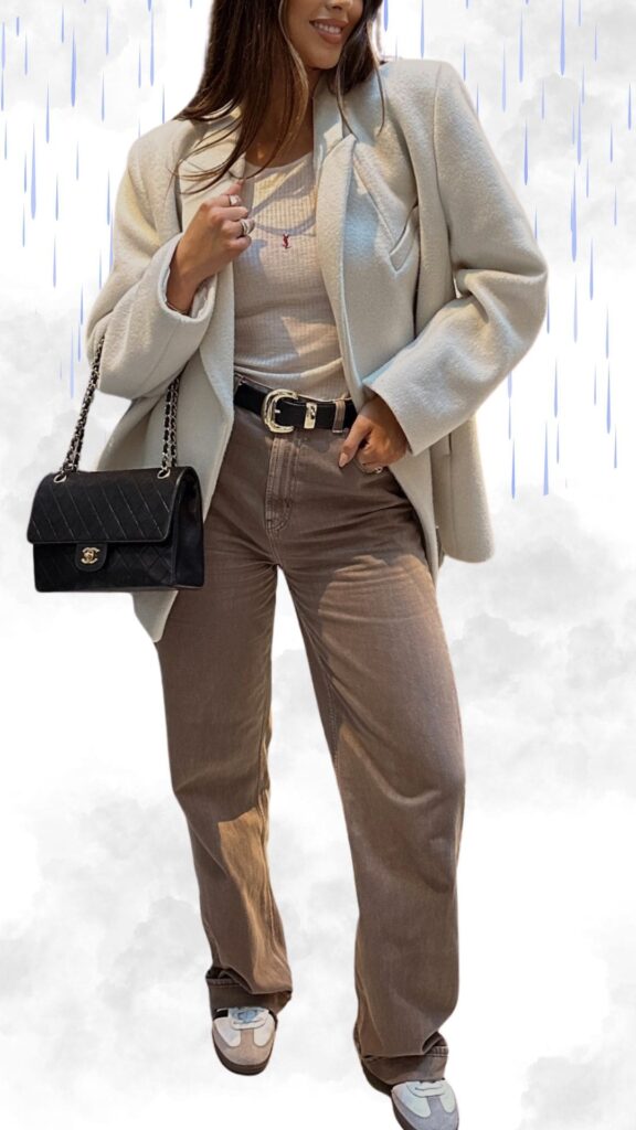 The outfit features a soft, cream oversized coat layered over a lightweight, ribbed white long-sleeve top, paired with taupe wide-leg pants that add a relaxed yet chic vibe. Accessories include a stylish black quilted handbag and a sleek black belt, while the footwear consists of trendy neutral-toned sneakers with subtle detailing for comfort and flair. This combination provides a fashionable yet practical look for rainy fall and winter days.