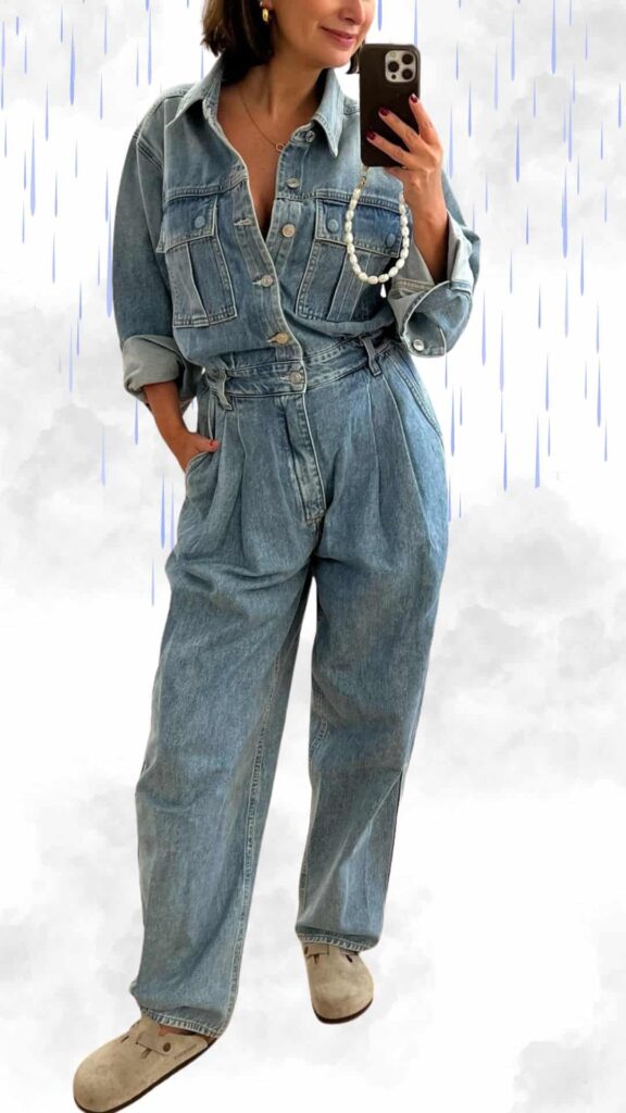 The outfit features a light blue denim jacket layered over a matching denim jumpsuit, showcasing a relaxed fit with pleated detailing for added style. The ensemble is completed with soft, beige slip-on clogs, offering comfort while maintaining a chic appearance, perfect for staying warm and dry on a rainy day. A pearl necklace adds a touch of elegance to this casual yet sophisticated look.