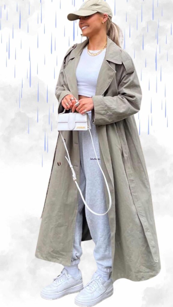 The outfit features a long, muted olive-green trench coat made from a lightweight, water-resistant fabric, layered over a fitted white crop top. Paired with soft gray joggers and white, chunky-soled sneakers, the look is both casual and chic, further complemented by a matching light-colored baseball cap and a small, structured handbag.