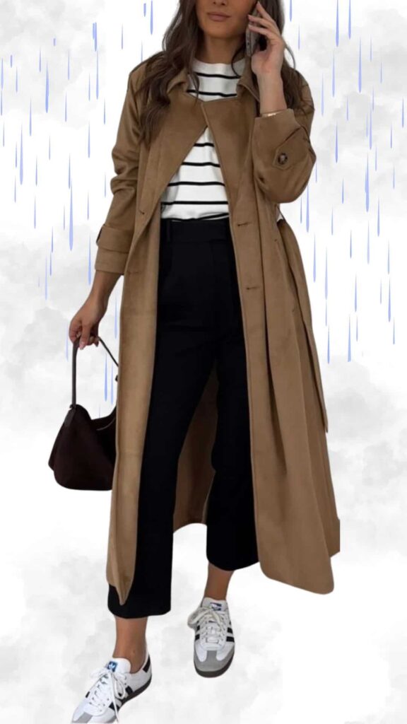 The outfit features a long, tan trench coat with a soft suede texture, layered over a classic white and black striped shirt. Paired with tailored black pants and white sneakers with gray accents, the look is both chic and practical, complemented by a dark brown handbag for added sophistication.