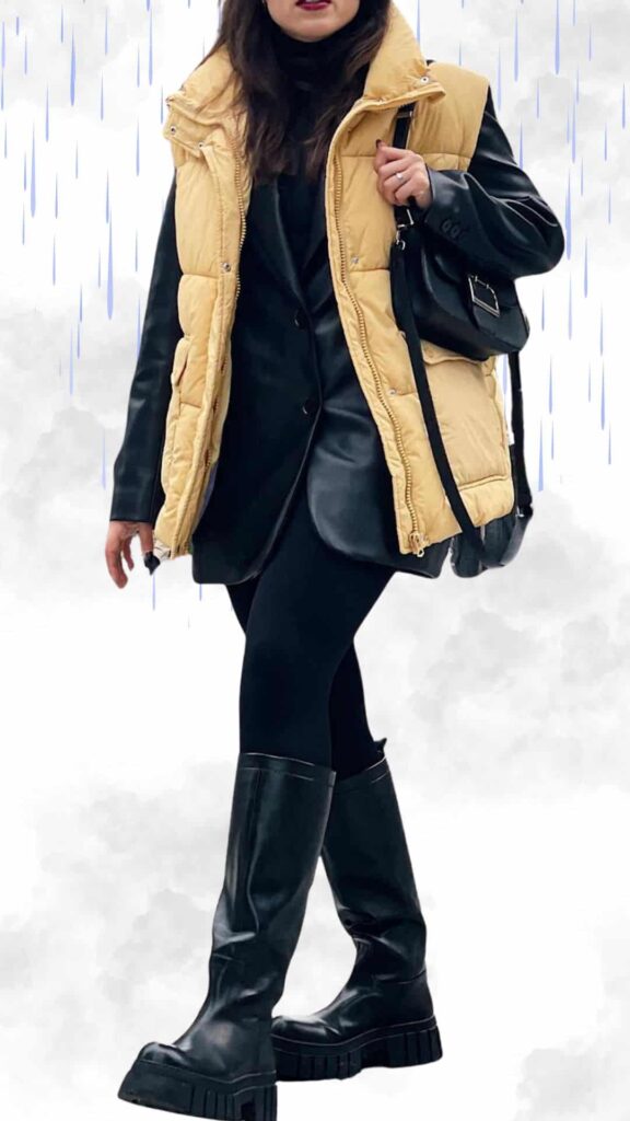 This chic rainy day outfit features a cozy, mustard yellow puffer vest layered over a sleek black leather blazer, paired with form-fitting black leggings. The ensemble is completed with stylish knee-high black platform boots, ensuring warmth and a fashionable look, while a small black handbag adds a touch of elegance. The combination of textures and bold colors creates a sophisticated yet practical choice for wet fall and winter weather.