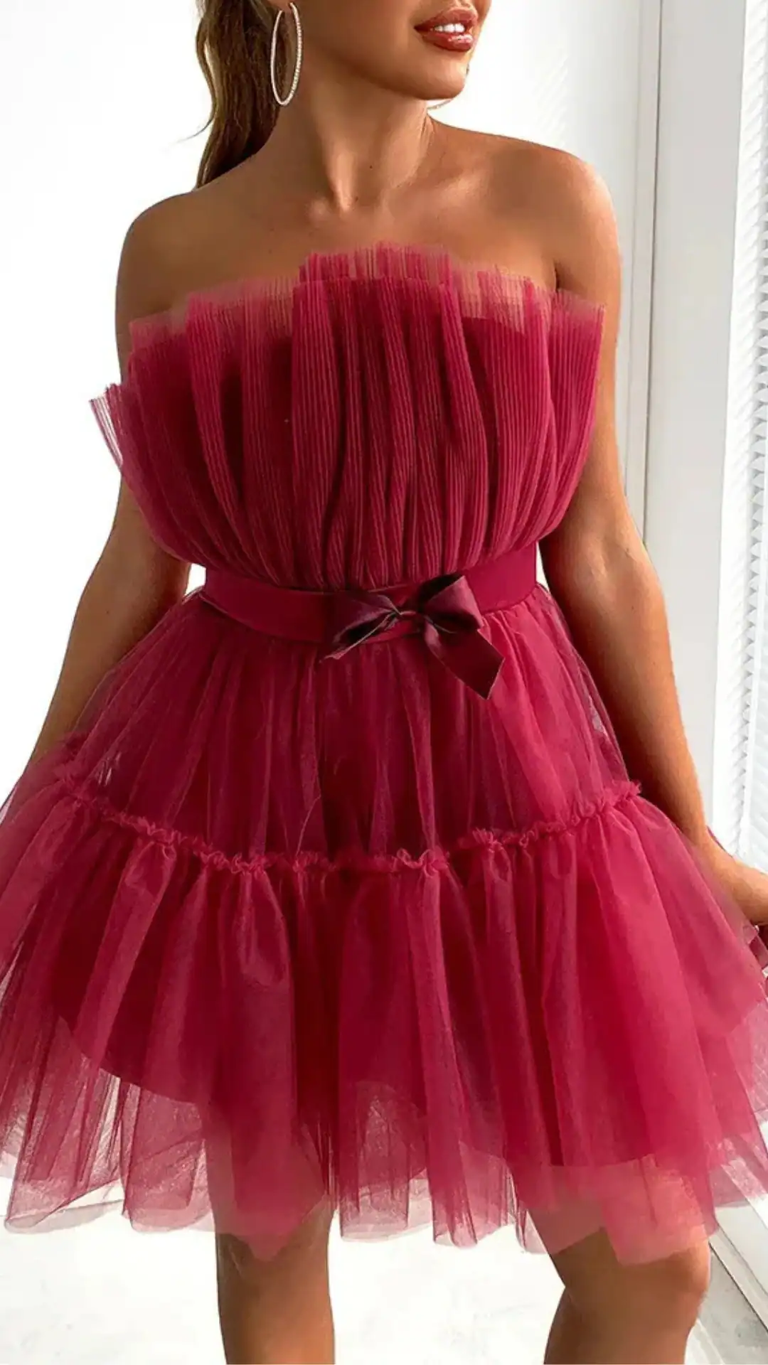 This red homecoming dress features a playful, strapless design with a fitted bodice made of pleated fabric, leading into a voluminous tulle skirt adorned with layered ruffles. Its vibrant berry hue and soft texture create a romantic and whimsical vibe, making it ideal for a festive school dance or party for teenagers.