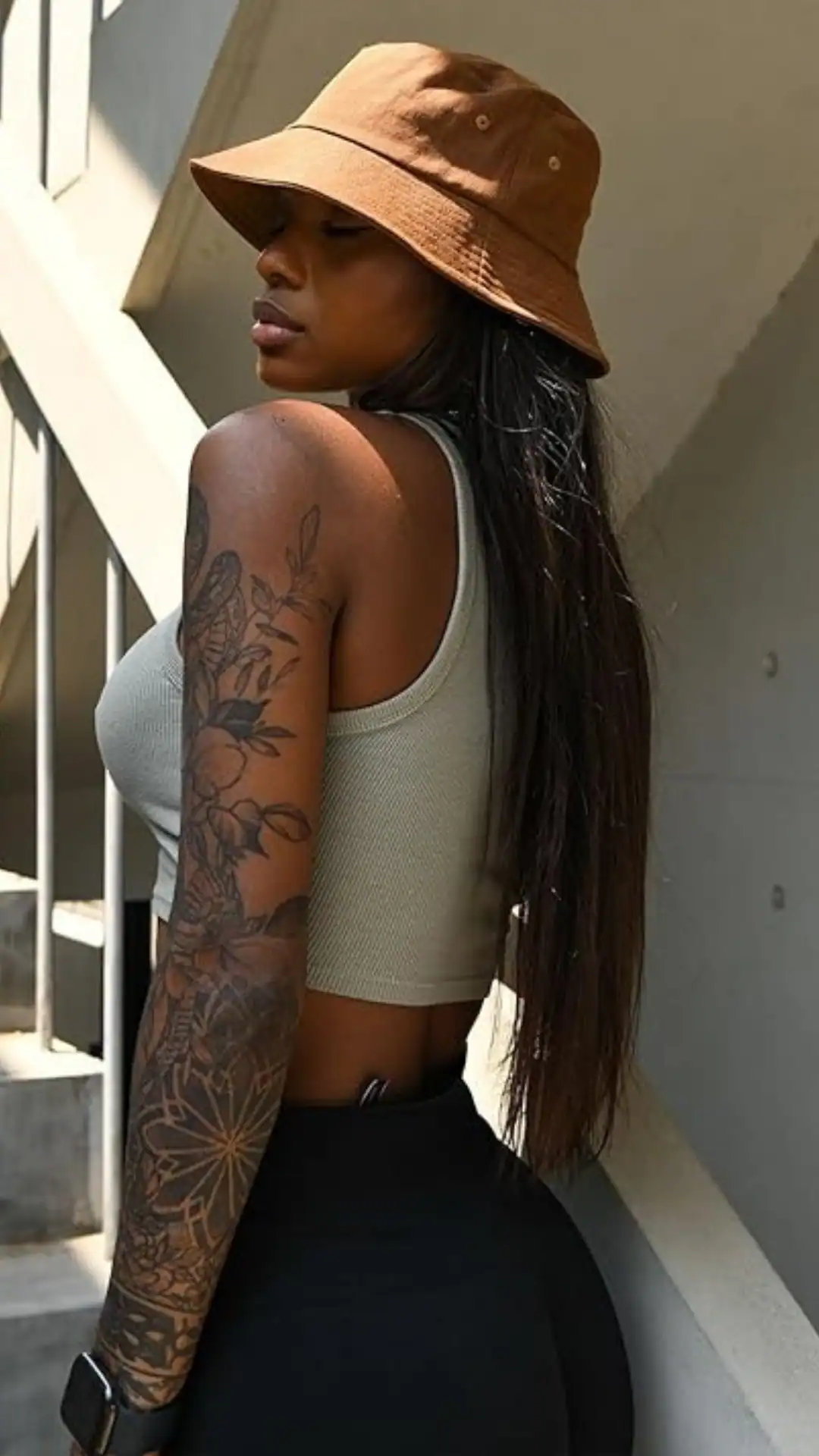 The outfit features a fitted, ribbed crop top in a soft sage green paired with high-waisted black leggings, creating a sleek, minimalist silhouette. Complemented by a classic brown bucket hat, this ensemble conveys a casual, urban vibe suitable for warm, rainy summer outings.