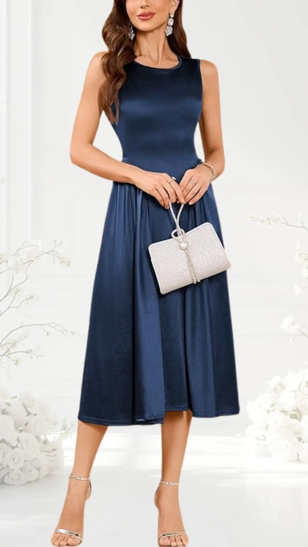 The dress is a navy blue, semi-formal sleeveless design that flows into a mid-length skirt. The model pairs it with metallic strappy sandals and holds a light silver, textured clutch. The backdrop features soft white florals, enhancing the elegant presentation.