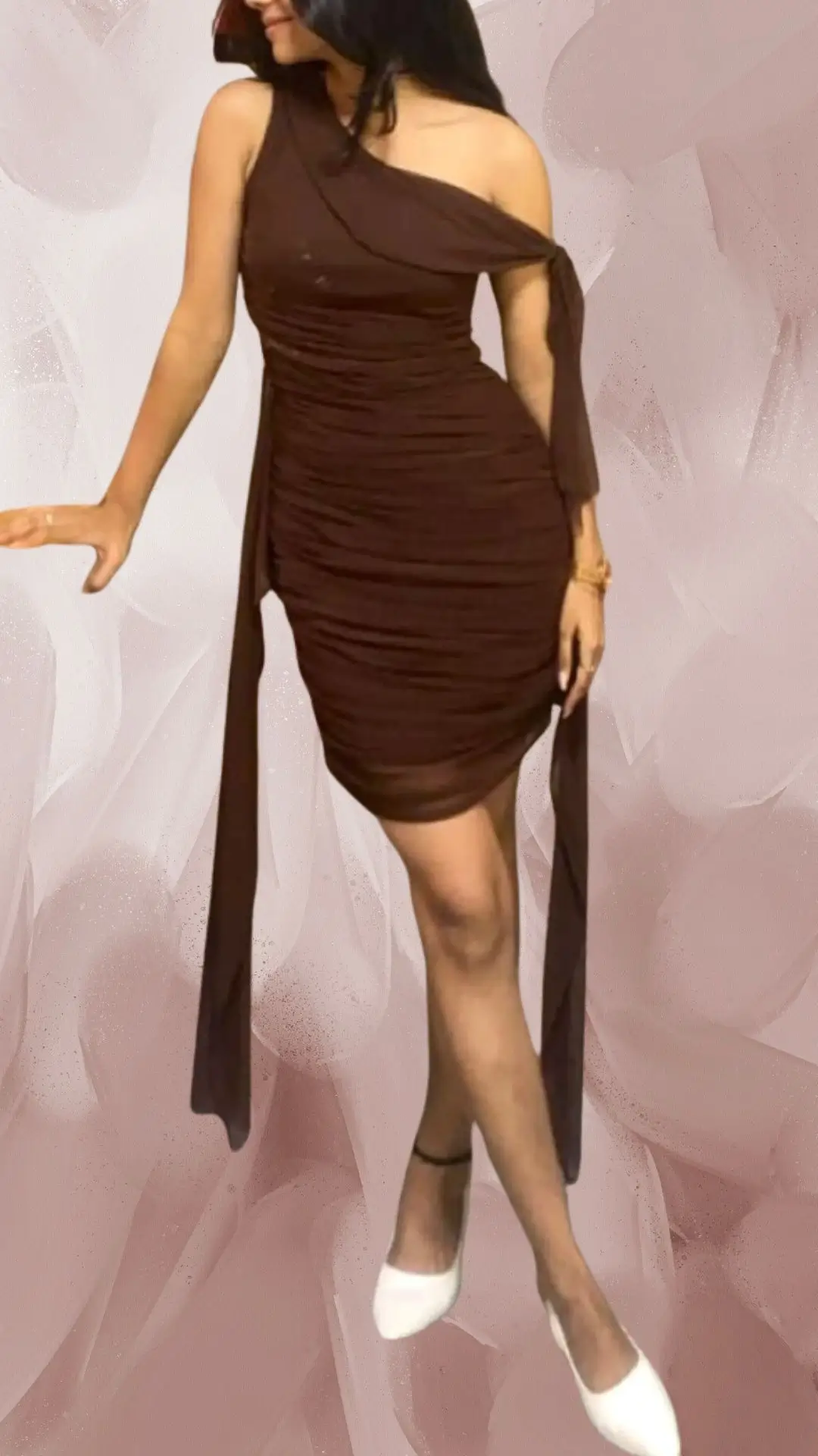 The outfit features a fitted, one-shoulder brown dress made from a soft, stretchy fabric, accentuated by ruching that sculpts the silhouette. The dress is paired with elegant white pointed-toe flats, adding a chic contrast to the deep brown color. A long, flowing fabric detail cascades from the waist, enhancing the sophisticated appearance of the ensemble.