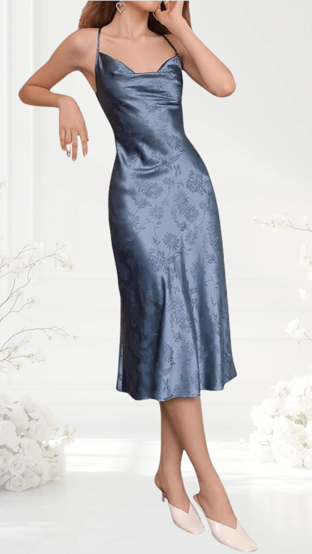 A satin slip dress in a muted blue hue features a fitted bodice with thin straps and a scooped neckline, elegantly cascading to a knee-length flared hem. The dress is adorned with a subtle floral pattern, enhancing its romantic appeal, complemented by beige pointed-toe heels. The background is softly blurred, highlighting the dress's design and texture.