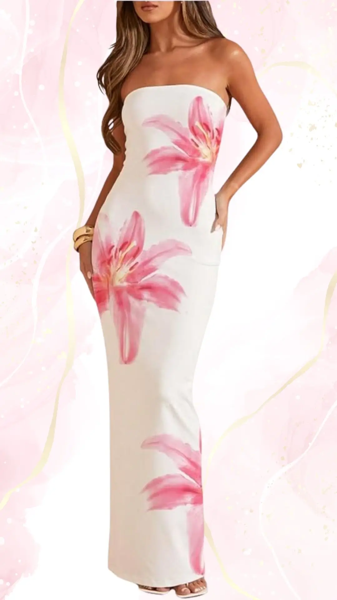 A strapless, form-fitting wedding guest dress features a soft white fabric adorned with large, hand-painted pink lilies. The floor-length design elegantly complements the figure, while the model poses with one hand in her pocket, showcasing a relaxed and stylish look.