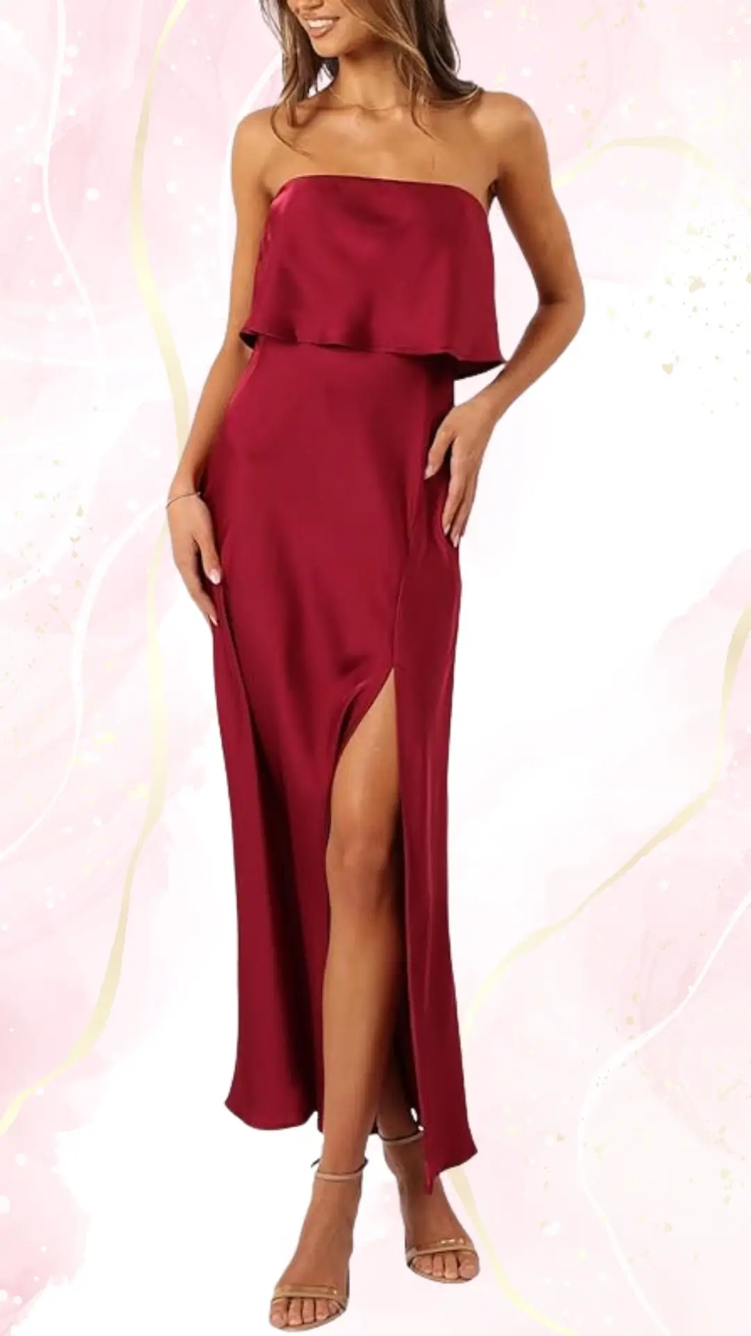 A summer strapless wedding guest dress in a deep burgundy shade features a layered bodice and a thigh-high slit for added elegance. The flowing fabric accentuates a sleek silhouette, while the model pairs it with delicate strappy sandals. The background is soft pink with subtle abstract designs, enhancing the dress's vibrant color.
