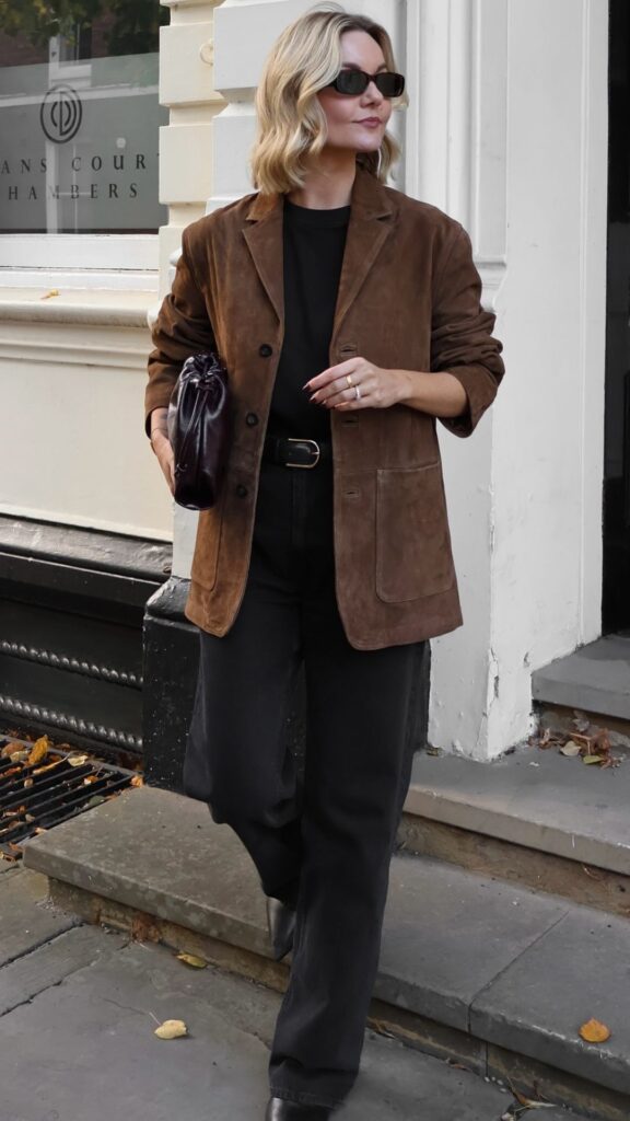 The outfit features a chic brown suede jacket, tailored with a single-button front and patch pockets, layered over a fitted black t-shirt. The look is completed with high-waisted black trousers that flare slightly at the bottom, creating a sophisticated silhouette, along with pointed-toe black ankle boots and a small, glossy dark purple handbag, adding a touch of elegance. The overall combination of textures and colors enhances the stylish appeal of the ensemble.