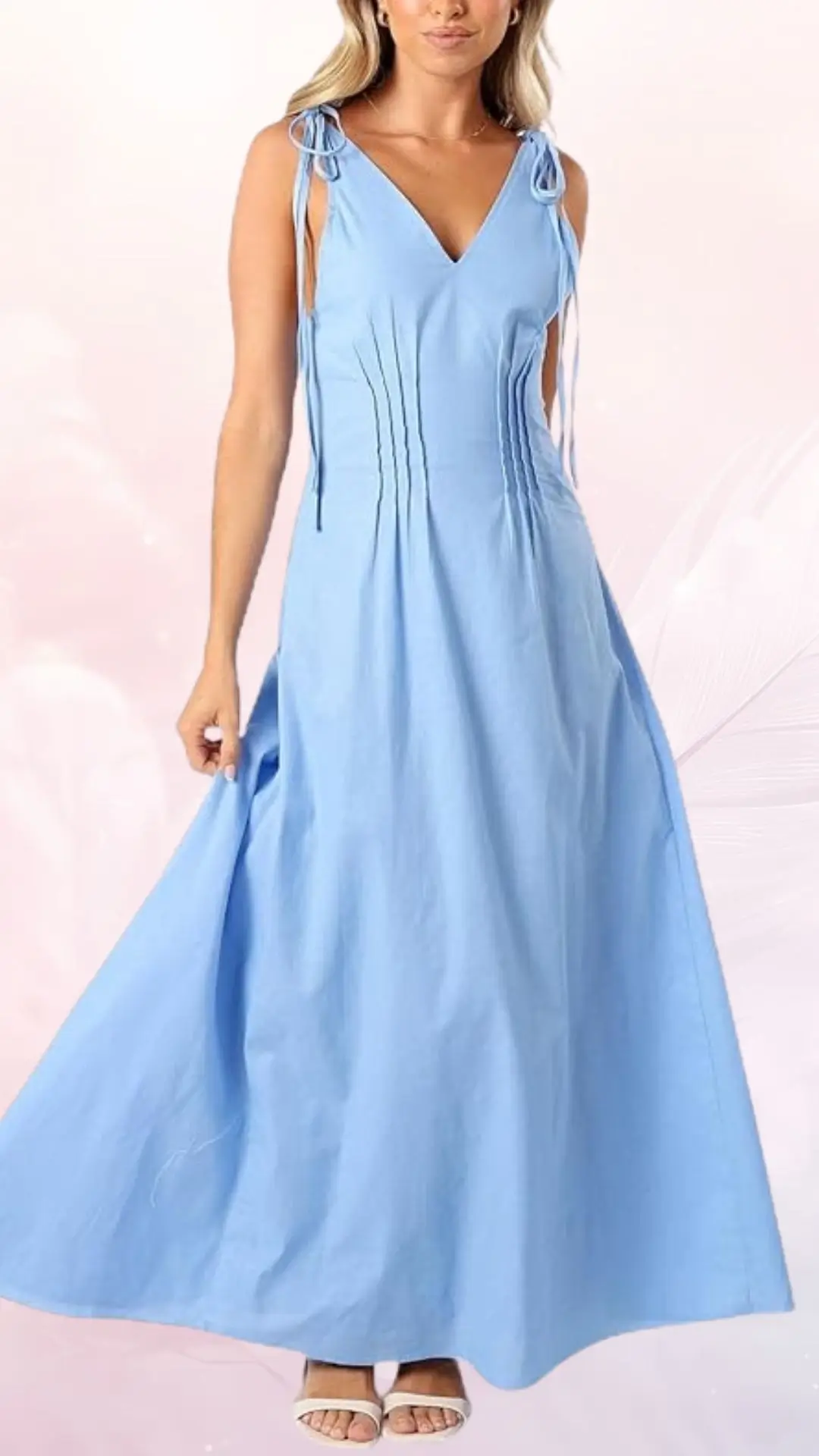 A lightweight, long dress in soft pastel blue features a deep V-neckline and delicate tie straps on the shoulders. The A-line silhouette flows gracefully, enhanced by subtle pleats along the bodice, creating an elegant and airy appearance, perfect for a summer wedding. The model pairs the dress with simple white sandals, emphasizing its casual yet sophisticated style.