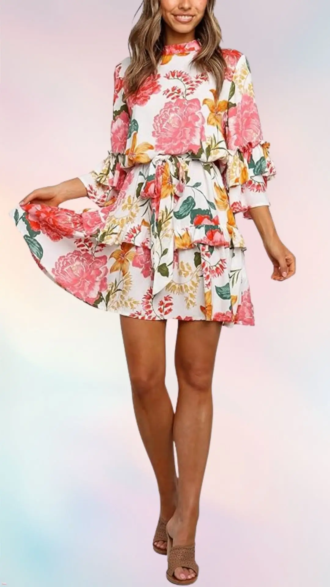 A floral summer dress features bold pink and orange flowers against a white background, with a ruffled hem and sleeves. It is cinched at the waist with a matching belt, creating a playful silhouette. The model pairs the dress with light tan heeled sandals.
