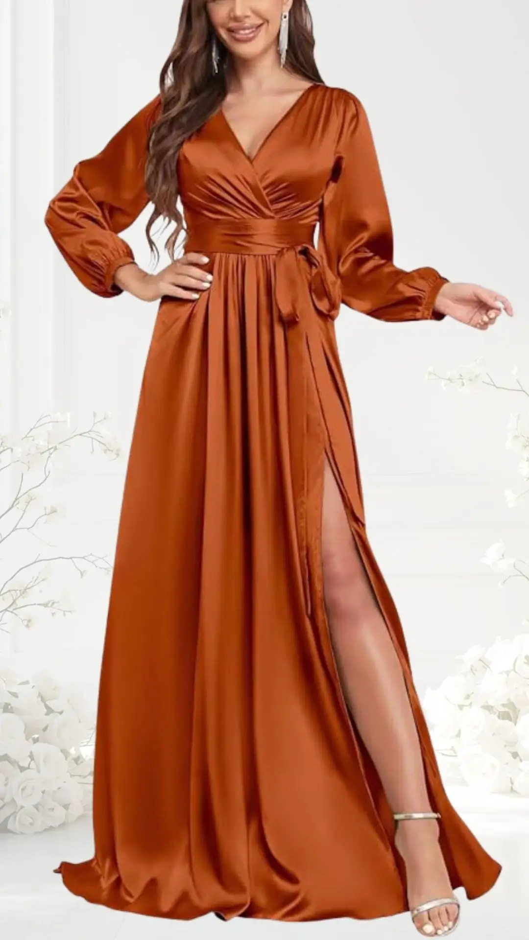 A terracotta wedding guest dress features a deep V-neckline and long, billowy sleeves, cinched at the waist with a fabric tie. The floor-length gown has a daring side slit, revealing a hint of leg, and is gracefully made from a silky material. The model complements the dress with strappy silver heels, set against a soft, neutral background with delicate floral accents.
