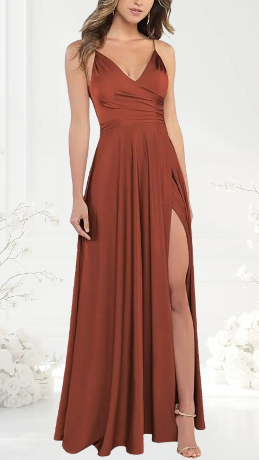 The terracotta wedding guest dress features a fitted bodice with thin spaghetti straps, flowing into a full skirt with a high side slit that reveals the leg. The smooth fabric drapes elegantly to the floor, complemented by delicate silver accessories. The background includes soft white floral accents, enhancing the dress's rich color.