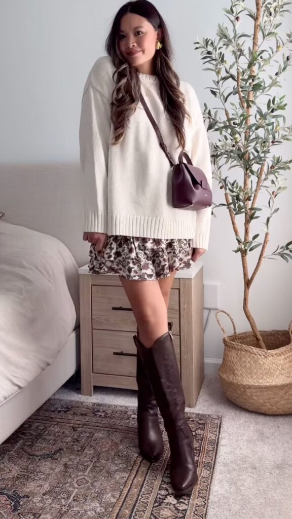 The outfit features an oversized, cream-colored knit sweater with a relaxed fit, layering over a flared mini skirt adorned with a delicate brown floral print, perfect for fall. Brown knee-high boots with a sleek finish complete the look, while a small, rich burgundy handbag adds a chic touch, enhancing the overall warm autumn aesthetic. The combination of textures and colors creates a cozy yet stylish ensemble ideal for Thanksgiving gatherings.