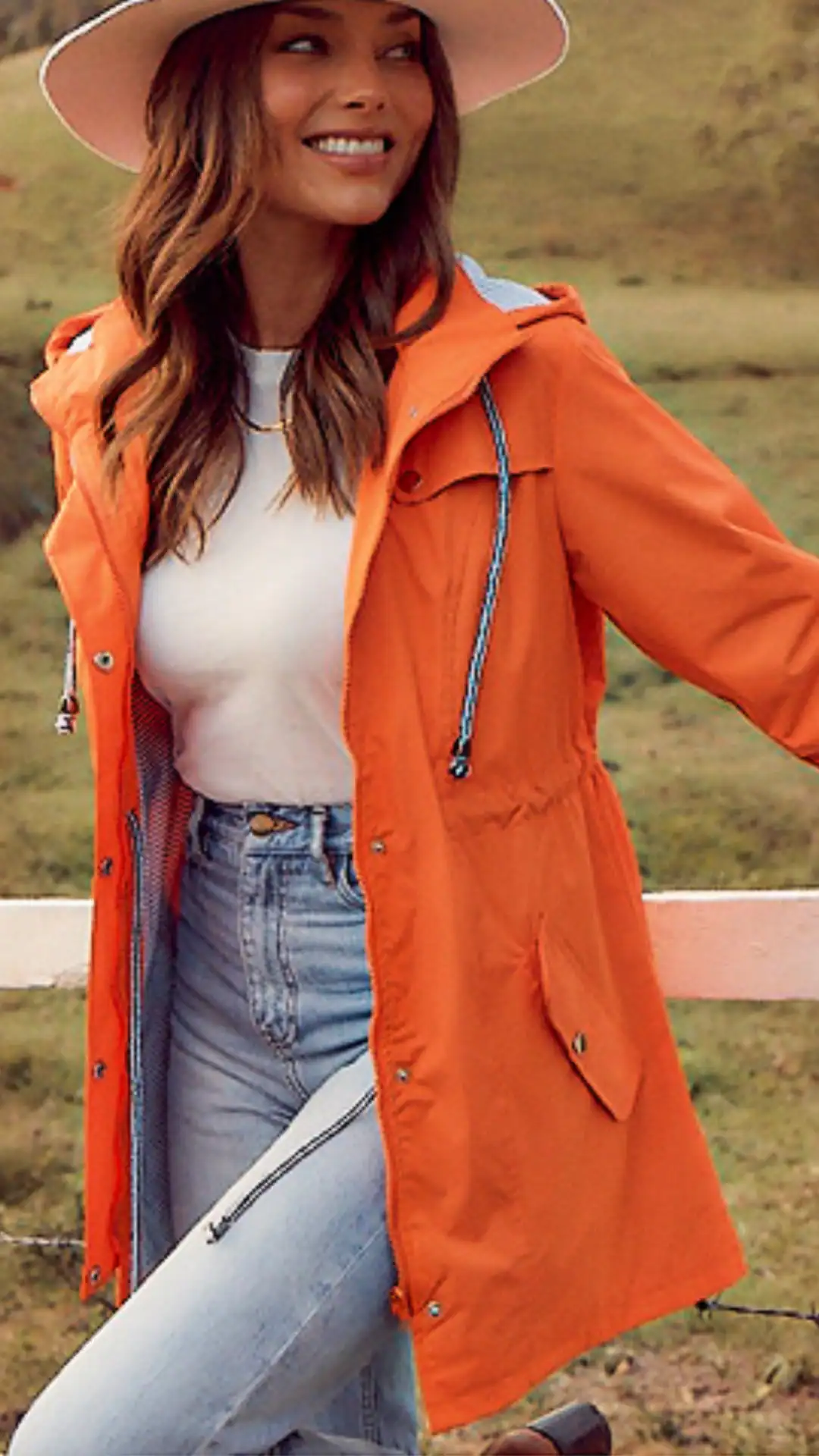This vibrant orange rain jacket features a lightweight, waterproof fabric with a smooth texture and adjustable drawstrings at the waist, paired with a classic white t-shirt and casual light-wash jeans, creating a fresh, playful look ideal for summer outings. The overall vibe is upbeat and modern, suitable for casual events or outdoor activities in rainy weather.