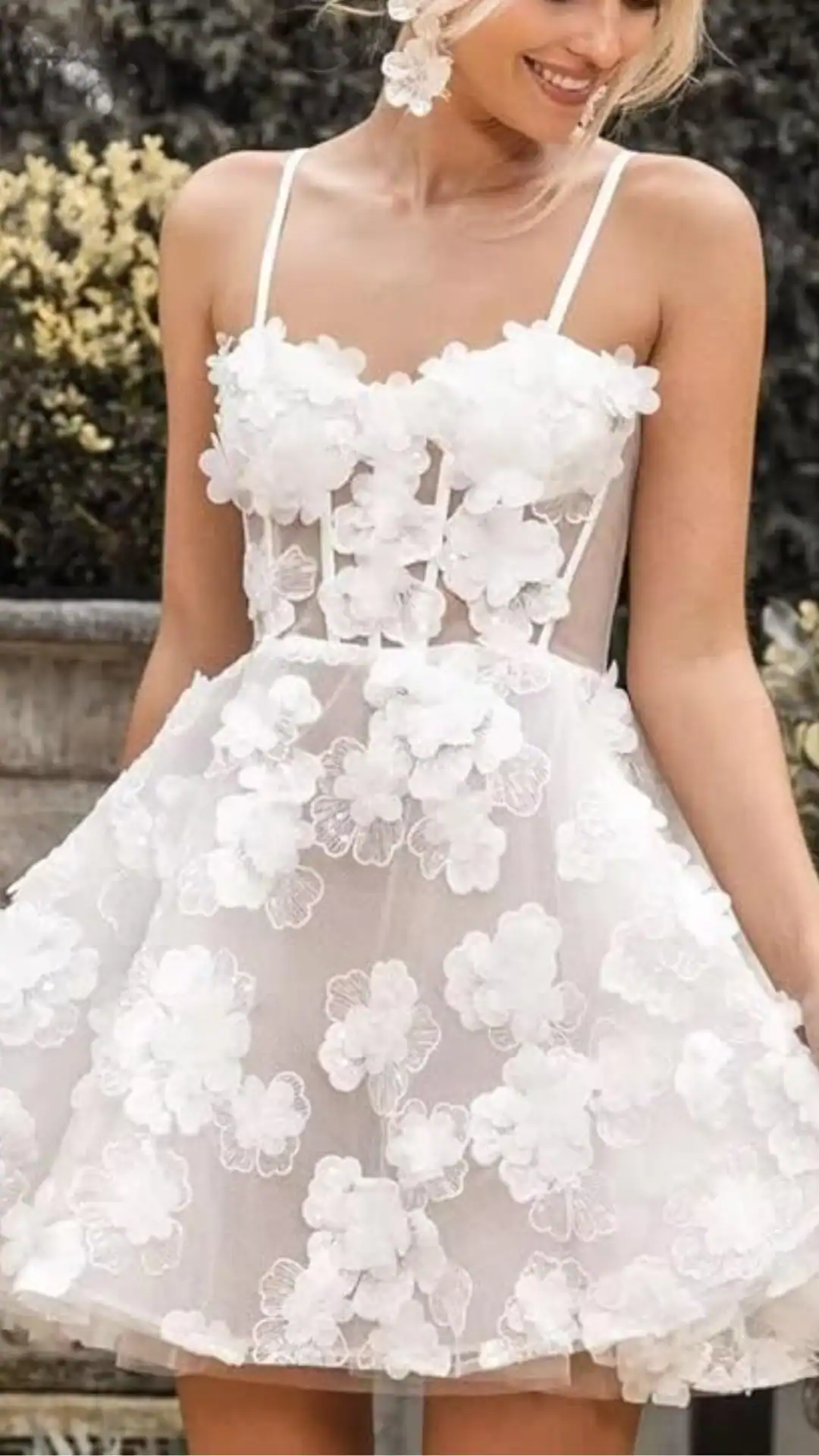 This whimsical white homecoming dress features a fitted, floral-embellished bodice with delicate straps and a voluminous tulle skirt, creating a playful and romantic silhouette. The dress, adorned with 3D floral appliqués, is perfect for teens attending formal events like homecoming or school dances, exuding an ethereal and trendy vibe.