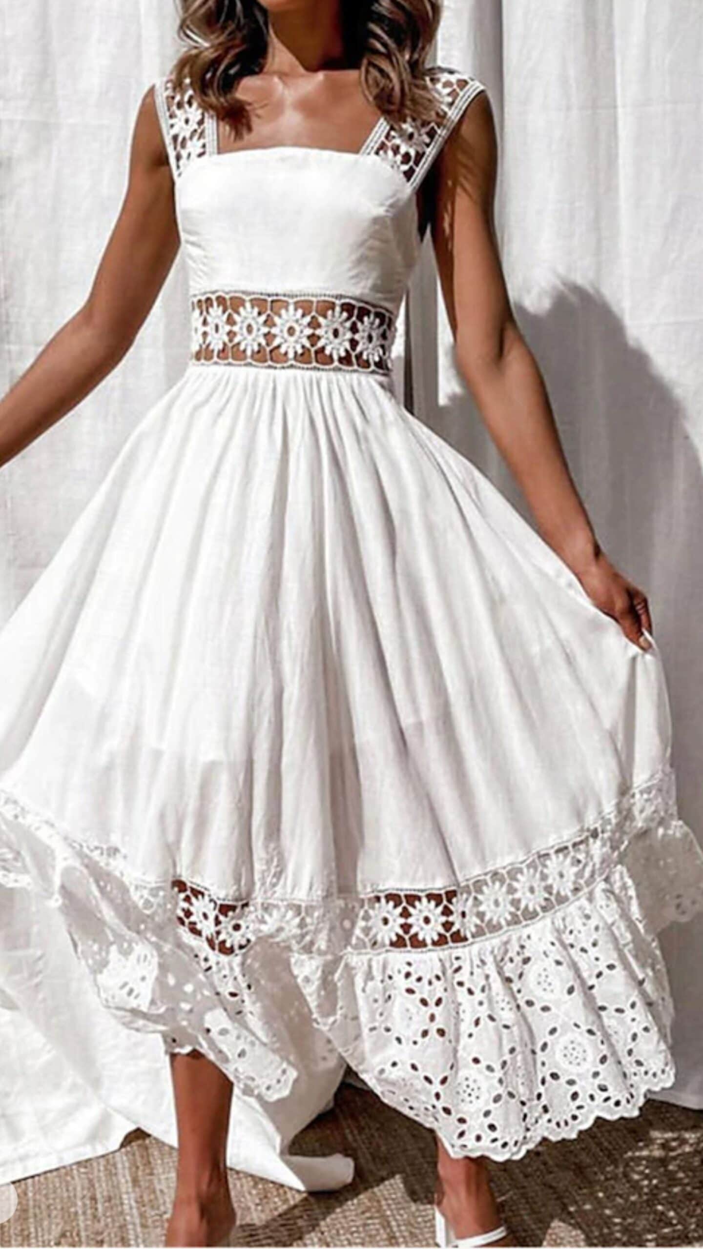 This white dress features a fitted bodice with a square neckline and wide straps, showcasing intricate floral lace trim along the waist and hem. Made from lightweight, textured fabric, it has a full, flowing midi length and is adorned with playful eyelet cutouts that contribute to a romantic and airy vibe, making it perfect for a casual summer outing or a garden party.