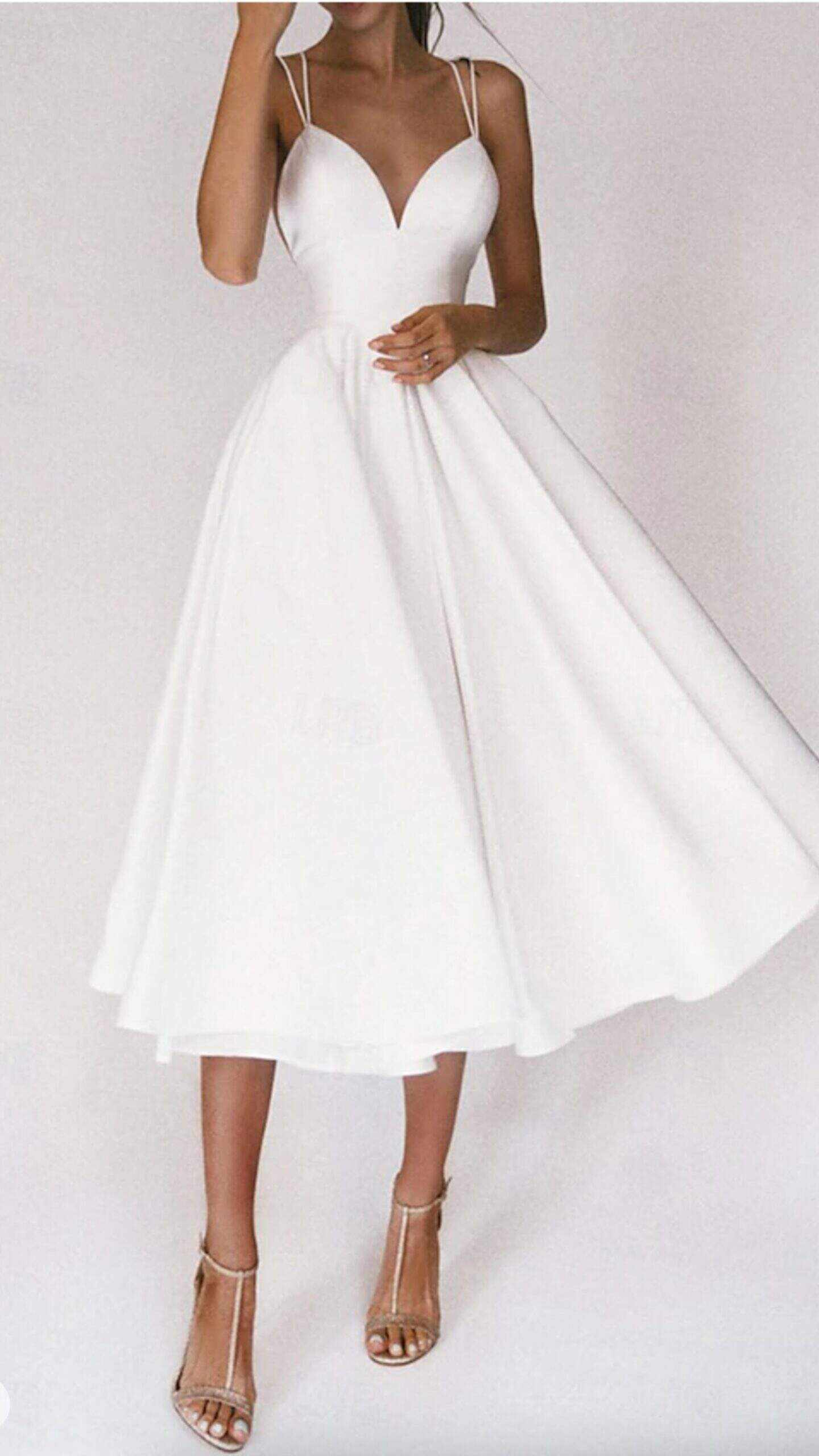 This white dress features a fitted bodice with a plunging V-neckline and delicate spaghetti straps, transitioning into a full, flowing skirt reaching a midi length. Made from a lightweight, matte fabric, it evokes a classic and romantic vibe, making it ideal for formal events or weddings, enhanced by the elegant movement of the skirt.