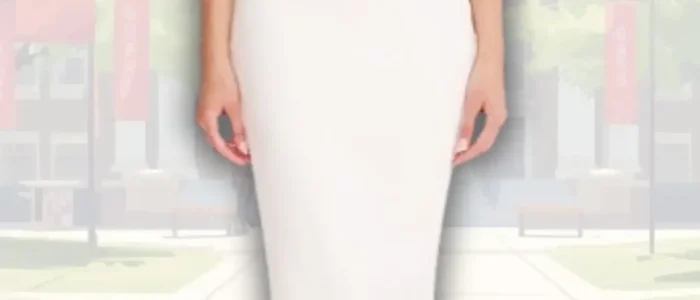 white-graduation-dress-with-asymmetrical-neckline-2025-12-14T01:17:18.000Z A fitted, sleeveless white graduation dress features a stylish asymmetrical neckline with a flowing shoulder detail. The dress falls to mid-calf, showcasing a sleek silhouette complemented by strappy nude heels. The background is a softly blurred outdoor setting with greenery.