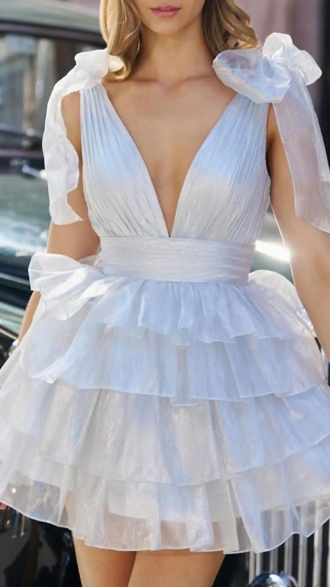 This enchanting white homecoming dress features a deep V-neckline and layered ruffles that create a playful, voluminous skirt, crafted from a shimmering, lightweight fabric that adds texture and movement. Embellished with oversized sheer bow accents on the shoulders, the overall vibe is romantic and whimsical, making it ideal for a teenage girl's special occasion such as homecoming or a school dance.