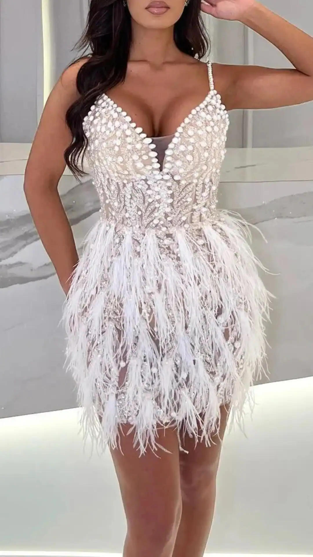 This white homecoming dress features a fitted bodice adorned with intricate beading and a plunging V-neckline, transitioning into a playful, feathered skirt that adds dynamic volume. The overall look exudes a bold, glamorous vibe, ideal for a festive occasion like homecoming, making it perfect for a teenage girl seeking a standout outfit.