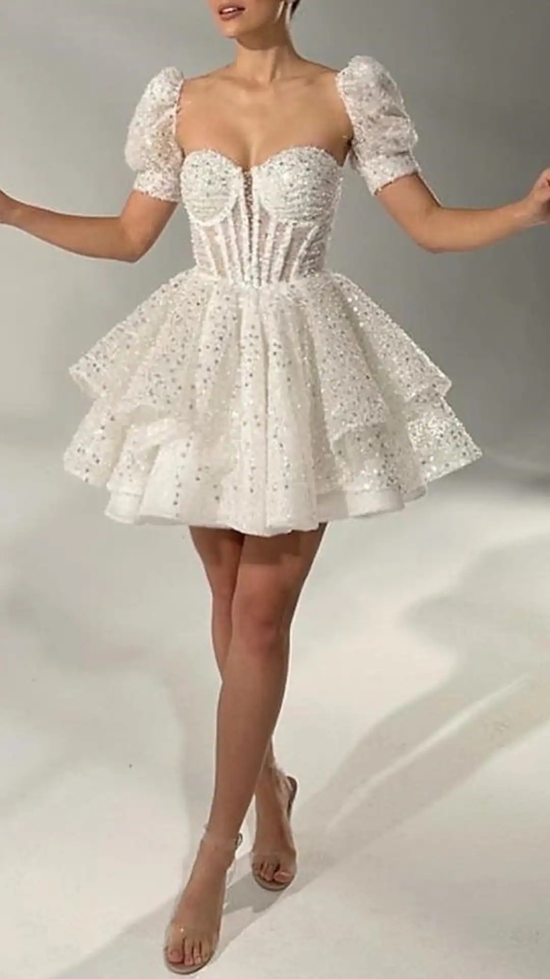 This white homecoming dress features a structured corset bodice with a sweetheart neckline, complemented by puffy sleeves and a layered, voluminous skirt adorned with delicate sequins, creating a dazzling effect. The overall vibe is whimsical and romantic, perfect for a formal event or school dance, designed specifically for teenage girls.