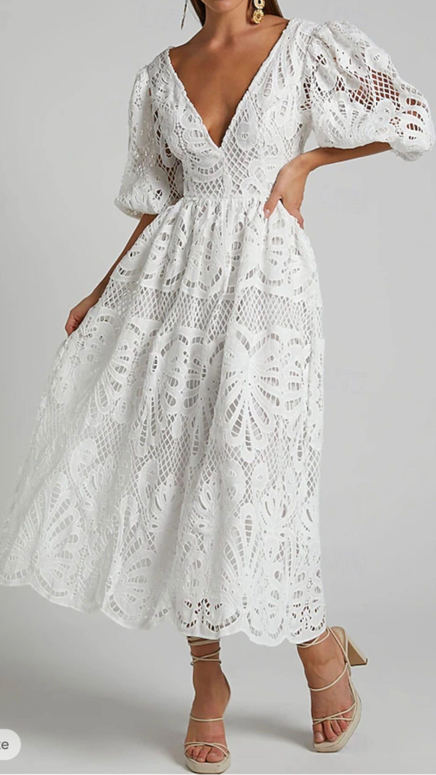 This white dress features a midi silhouette with a plunging V-neckline and billowy, puffed sleeves. Crafted from intricately embroidered lace, it has a textured, airy feel and showcases a floral pattern with cut-out details, exuding a romantic and feminine vibe, perfect for a garden wedding or brunch event.