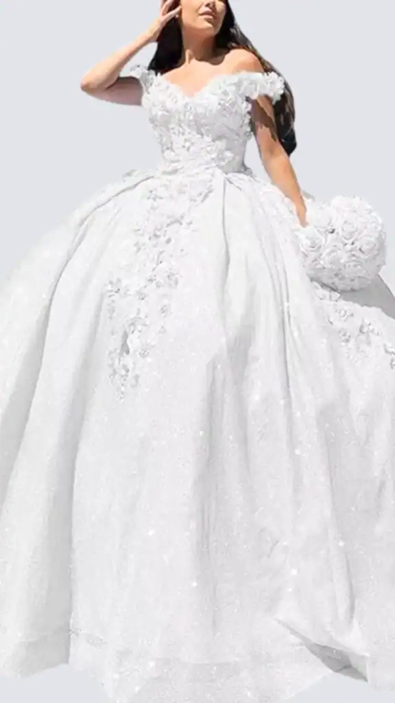 The dress features a fitted bodice adorned with intricate floral appliqués and delicate off-the-shoulder sleeves, creating an elegant and romantic look. The dramatic, voluminous skirt flows gracefully, made of shimmering white fabric that catches the light, while additional embellishments add a touch of sparkle. The overall silhouette accentuates the waist, making it ideal for a Quinceañera celebration.