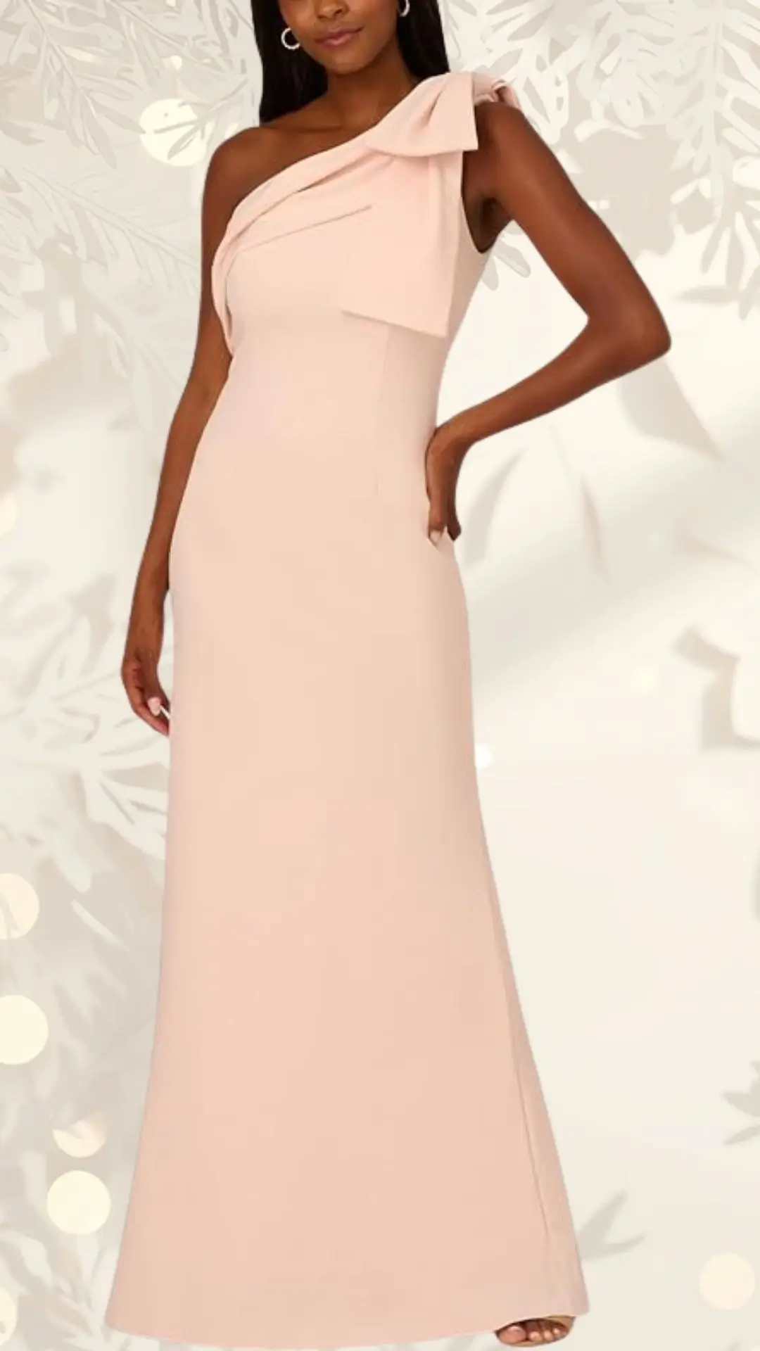 A slim, floor-length wedding guest dress in soft blush features a one-shoulder design accentuated by a draped bow at the shoulder. The fabric is smooth and form-fitting, creating an elegant silhouette that flows gently to the ground. The dress is styled against a subtle background with light, decorative elements.