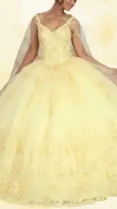 This yellow quinceañera dress features a fitted bodice with a deep V-neckline and delicate, sheer straps. The voluminous ball gown skirt is richly layered with sparkling embellishments and intricate floral appliqués, creating a whimsical look that flows gracefully to the floor. The overall shade is a soft, pastel yellow with subtle variations, enhancing the dress's romantic and elegant aesthetic.